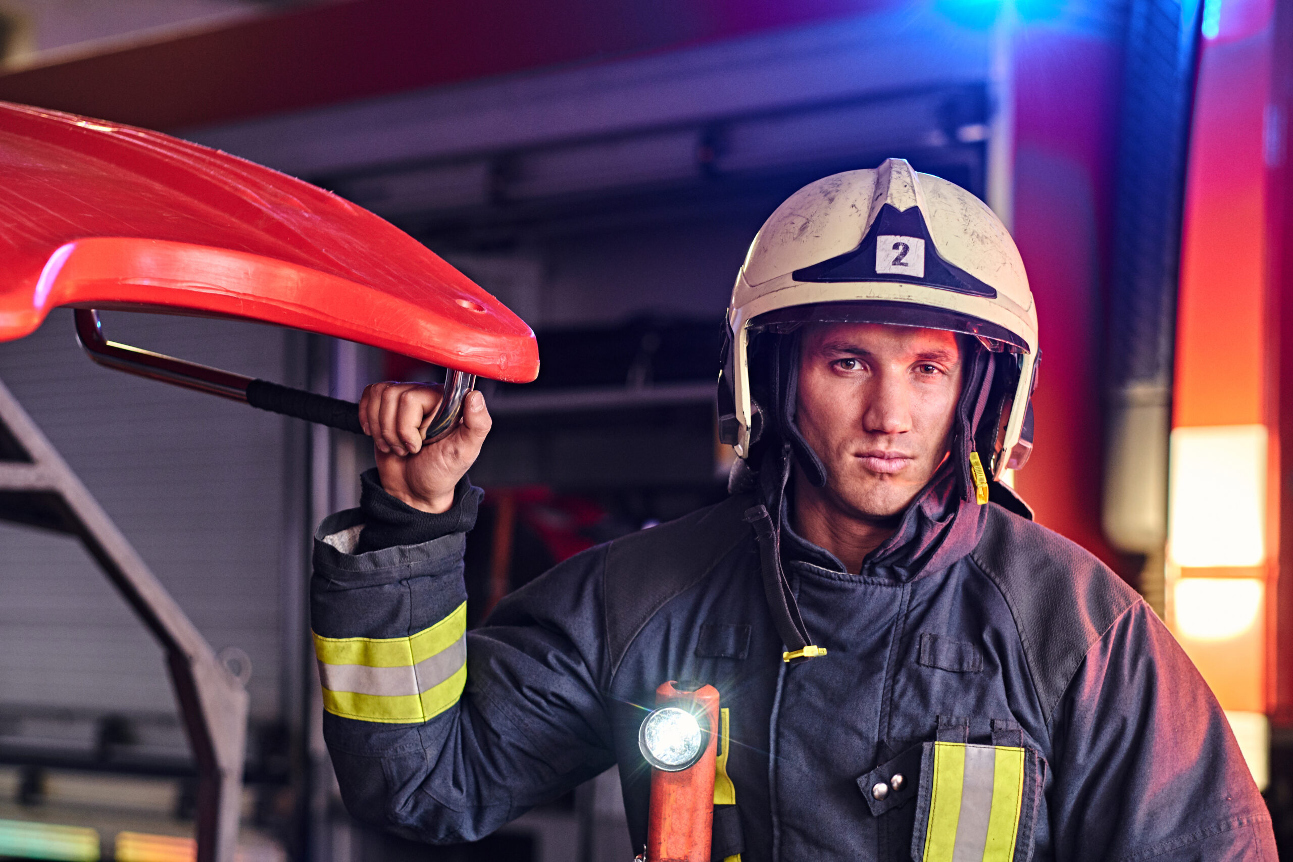 Portrait of a handsome fireman wearing a protective uniform with flashlight included standing in a fire station garage and looking at a camera